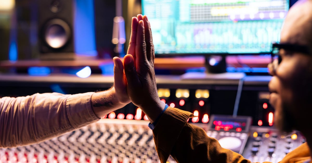 A close-up of two people's hands in a high-five gesture in front of a mixing console and a computer screen displaying audio software.